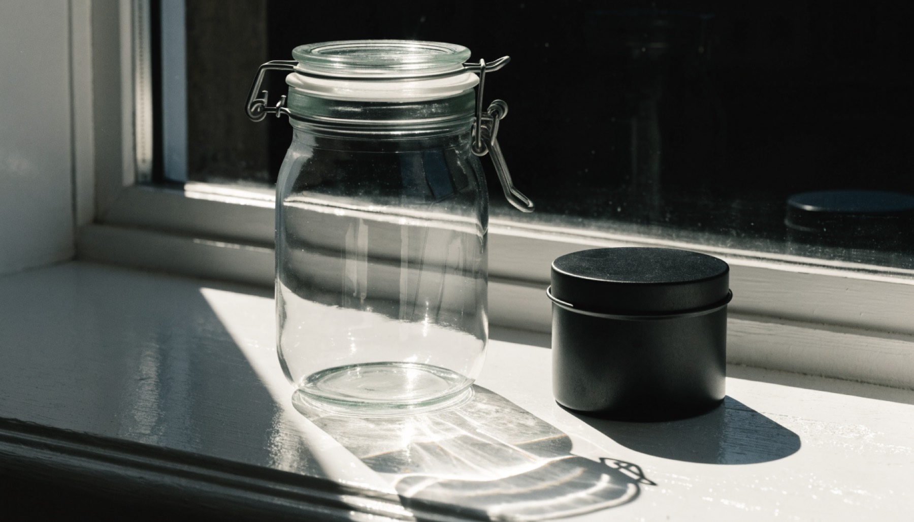 Un vaso di vetro trasparente vuoto su un davanzale bianco in pieno sole, con accanto una latta nera chiusa